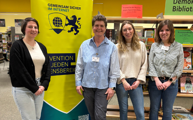 Vier Personen stehen in einer Bibliothek vor einem gelb‑grünen Banner mit der Aufschrift ‘Gemeinsam sicher im Internet’.