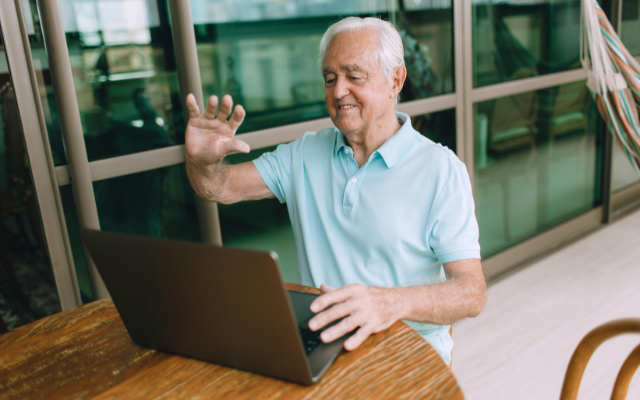 Eine ältere Person sitzt an einem Tisch vor einem Laptop und hebt zur Begrüßung die Hand