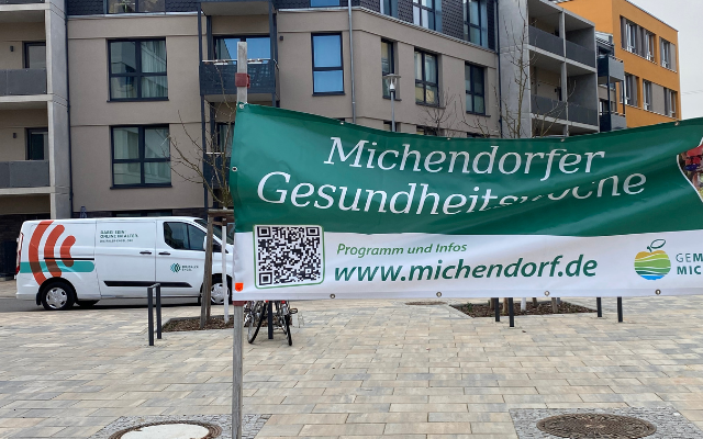Ein grünes Banner mit der Aufschrift ‚Michendorfer Gesundheitswoche‘ hängt auf einem Platz. Es zeigt einen QR‑Code sowie den Hinweis auf Programm und Infos unter www.michendorf.de. Im Hintergrund steht das weiß‑grüne Infomobil des Digitalen Engels, daneben moderne Wohnhäuser.